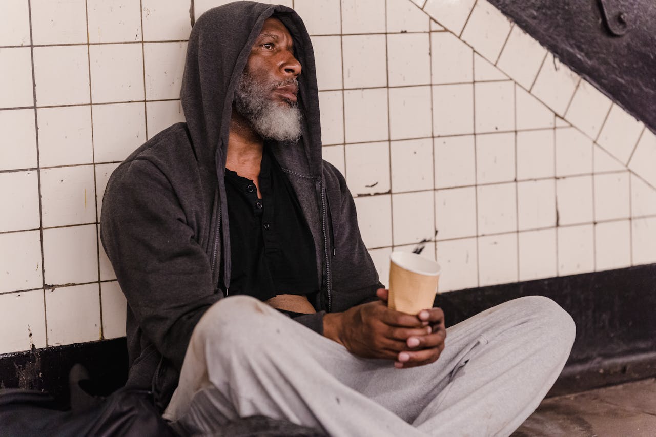 A man in a hoodie sits in a tiled corridor, holding a paper cup, conveying urban hardship.