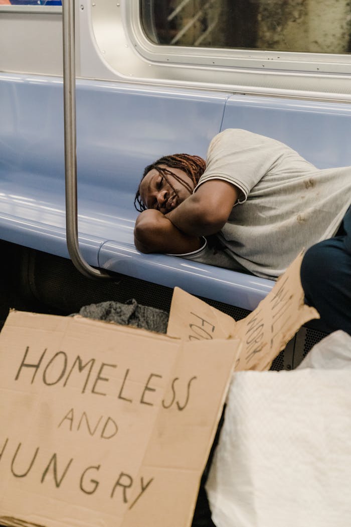 An African American homeless man is sleeping on a subway bench next to cardboard signs.