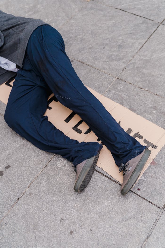 A person in blue pants lies on a sidewalk with a cardboard sign, symbolizing homelessness.